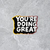 You're doing great! A vinyl sticker with white block lettering and black drop shadow, with eggplant to mustard yellow gradient around the edges. Hand lettered by Em Dash Paper Co. in Winston-Salem, NC.